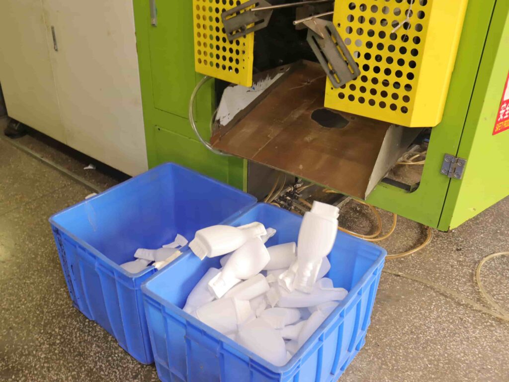 Blow molding machine discharging finished white cosmetic plastic bottles into blue plastic turnover bins in factory workshop